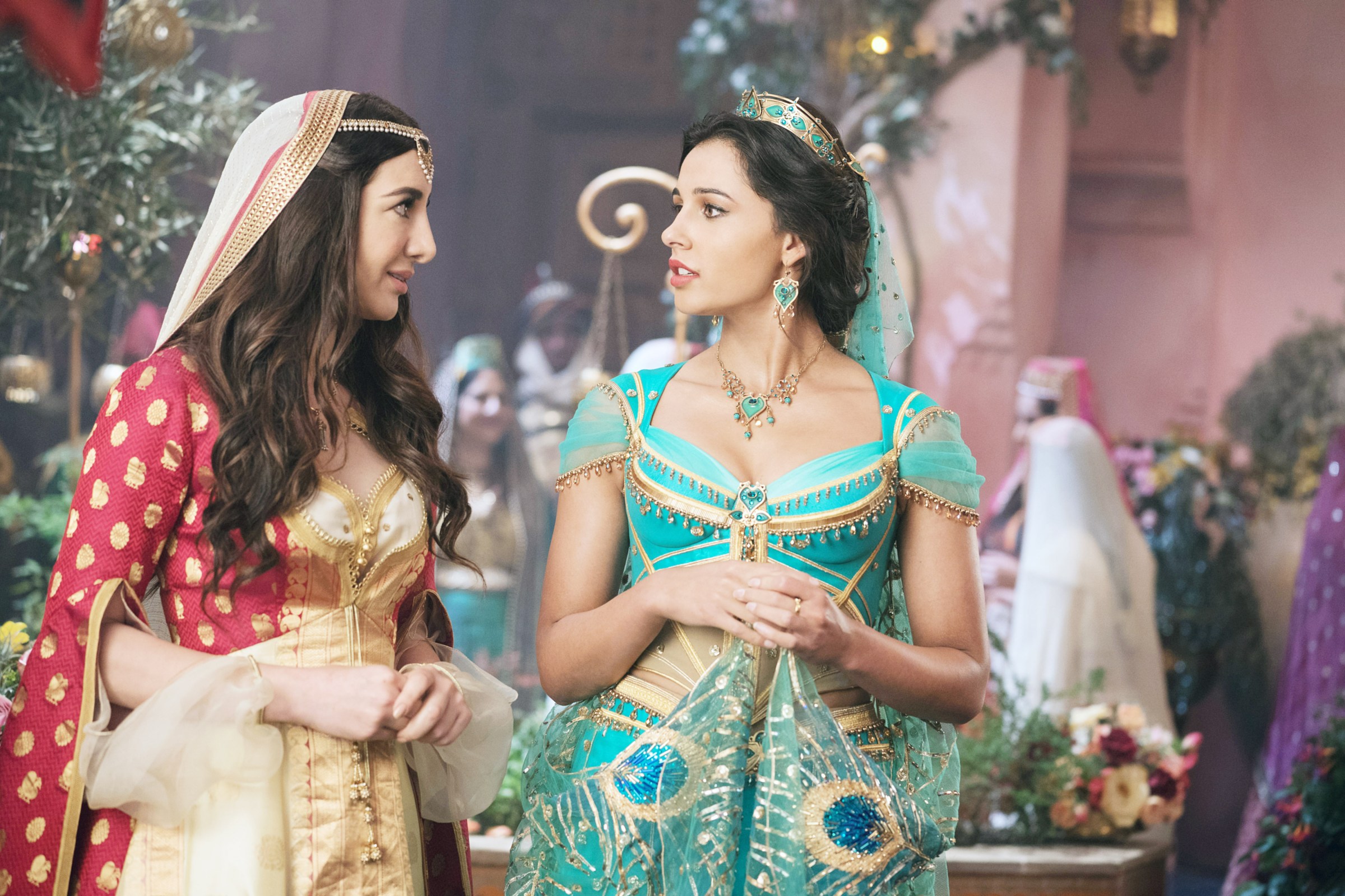 Jasmine (Naomi Scott) wearing a brightly colored turquoise outfit with peacock-feather beaded designs, with her handmaiden Dalia next to her. From Disney’s live-action Aladdin movie.