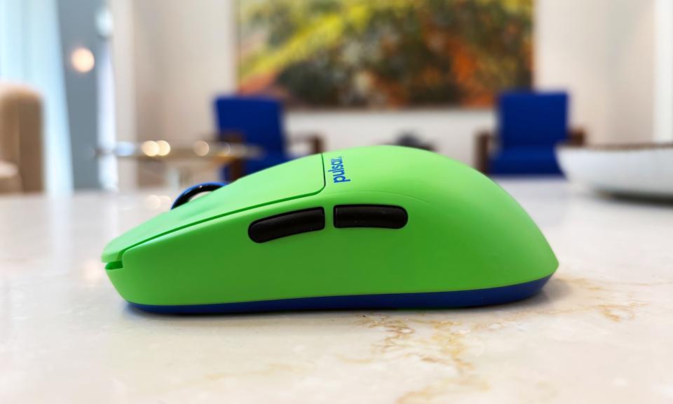 A lime green gaming mouse rests on a marble table in front of blue chairs. The mouse is shot from the side to display its rounded profile.