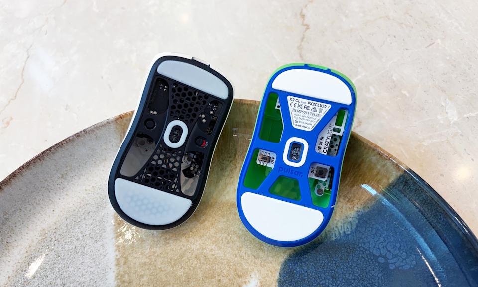 Two gaming mice rest in a brown and blue platter on a marble table, showing the cutouts on the bottom of their shells.