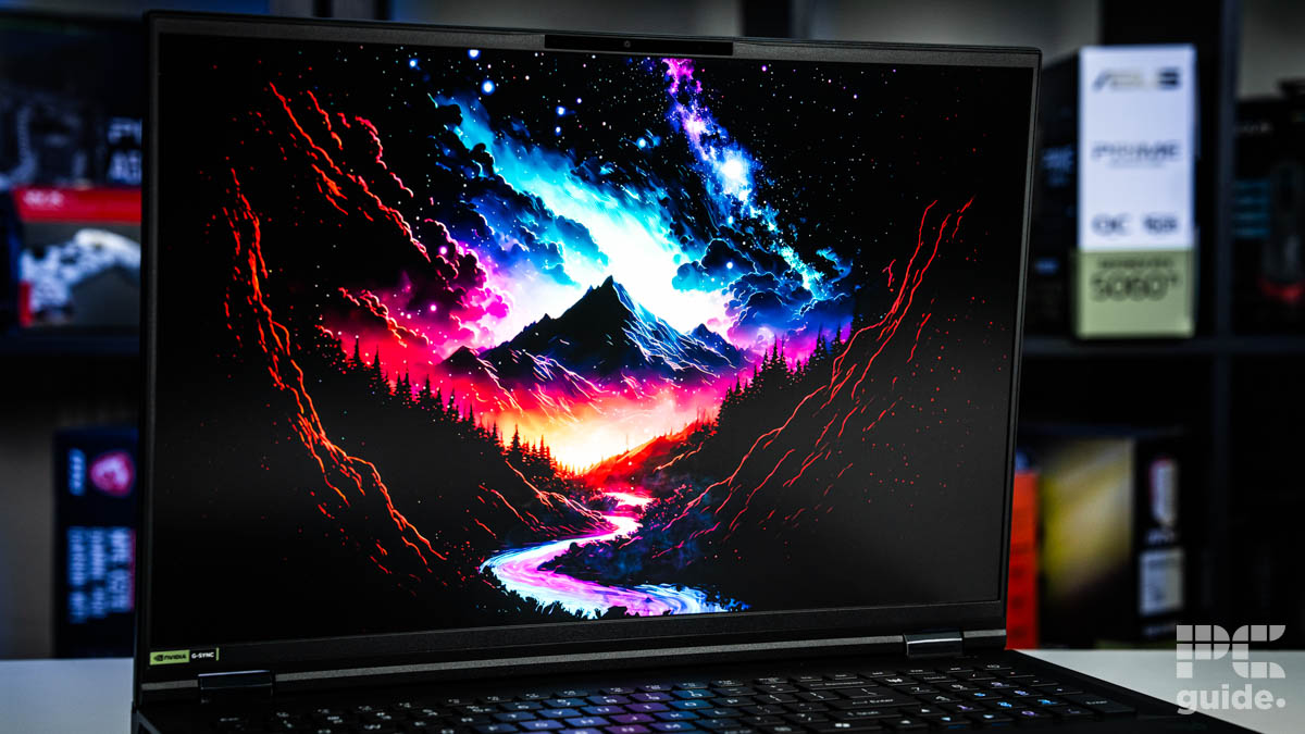 The screen of the laptop. it has a black mountain in the middle with black clouds and stars above and beyond. Around it, it glows in blue and pink that steams off above, with a river meandering towards the camera. Surrounded by trees and mountains ranging up. Sitting on a white table with a blurred background of shelves filled with boxed of tech products.