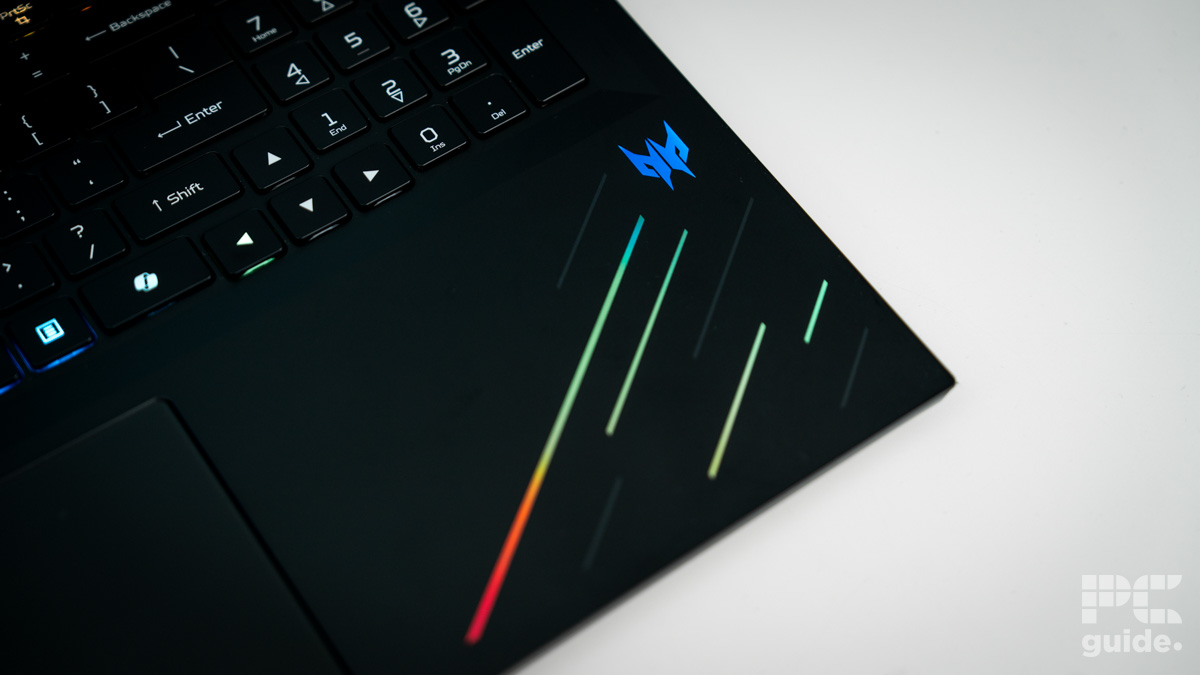 The bottom right edge of the laptop keyboard. With black base color, a predator logo in blue, and a few strips of RGB lights currently in green and red. With it sitting on a white table.