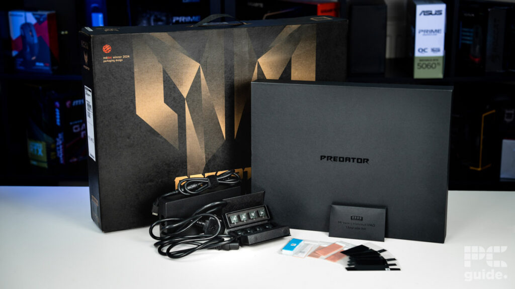 The boxes and contents of the laptop. On the back left is the main box in black witha bronze predator logo. On the right is a black laptop box with predator written in the middle. Bottom left is the power adapter and cable. In the foreground are the replacement keycaps and thermal upgrade. Sitting on a white table with a blurred background of shelves filled with boxed of tech products.