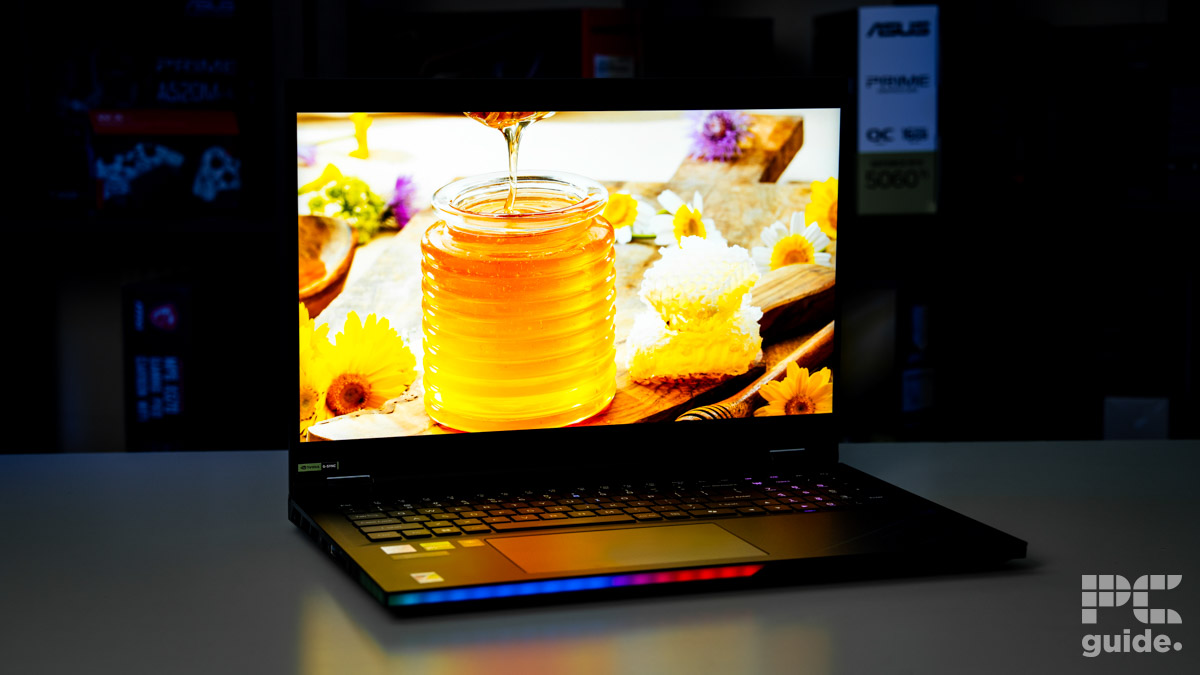 Black laptop open with RGB on the front edge, on screen is a very brright image of a glass jar filled with honey as some oozes off a stick from above. Sitting on a white table with a blurred background of shelves filled with boxed of tech products.