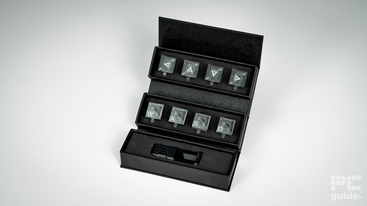 A set of replacement keycaps for the arrows and WASD, along with a keycap rremover tool. They sit in a fold out box that's on top of a white table.