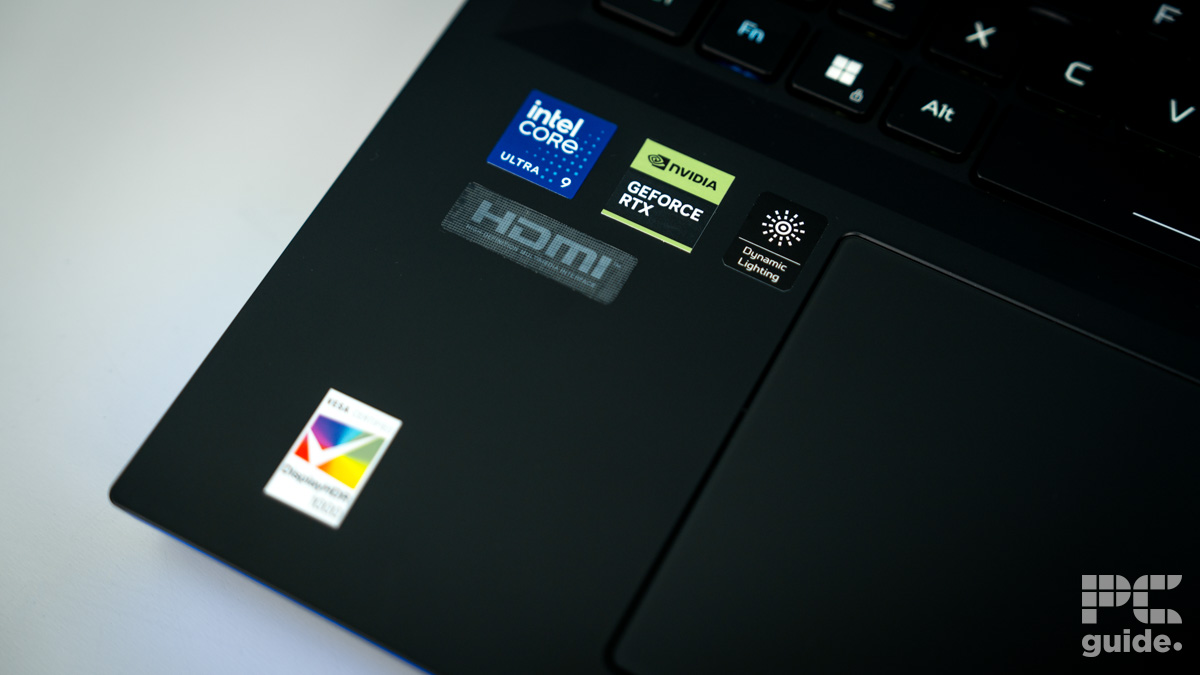 The stickers on the bottom left of the black laptop body. There is a blue Intel Core Ultra 9, Green and black Nvidia GeForce, White on black Dynamic lighting, a grey HDMI, and an out of focus white tick HDR. It sits on a white table.
