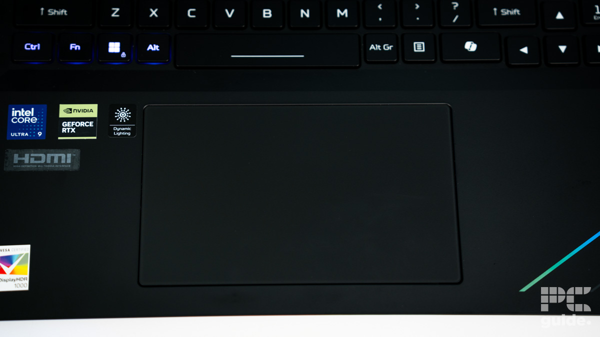 The black bottom edge of the laptop. In the middle is an ingressed touchpad. Stickers are on the left, keyboard bottom rows on the top.