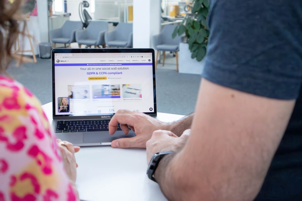 Two people sit at a table looking at a laptop screen showing the Walls.io homepage. The screen highlights GDPR & CCPA compliance and the option to create a free social wall.
